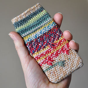 Une main tient une coque de téléphone tricotée, ornée de rayures horizontales colorées et de motifs dans des tons de vert, rouge, jaune et beige. L'arrière-plan est uni et neutre.