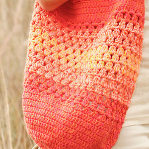 Gros plan sur une main tenant un sac au crochet dans des tons d'orange, de rose et de jaune, mettant en valeur son motif texturé. L'arrière-plan est flou, avec de hautes herbes.
