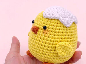 Une main tient un adorable poussin au crochet — un poussin jaune au petit bec orange, aux ailes minuscules, aux yeux noirs et coiffé d'une coquille d'œuf blanche au crochet — sur fond rose pâle.
