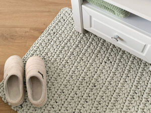 Des chaussons beiges sont posés sur un tapis gris à grosses mailles, qui rappelle un tapis au crochet, à côté d'une table de chevet blanche dotée d'un tiroir, sur l'étagère de laquelle on aperçoit une serviette pliée. On distingue également un parquet en bois clair.