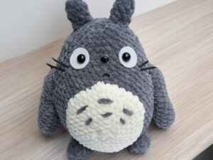 Une peluche ressemblant à Totoro, avec une fourrure grise texturée, de grands yeux ronds et un ventre blanc parsemé de taches grises, est posée sur une surface en bois clair ; son charme artisanal rappelle les créations « Totoro au crochet ».