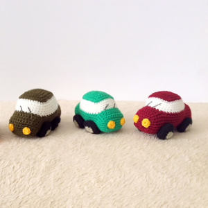 Trois petites voitures jouets au crochet — marron, verte et rouge — sont alignées côte à côte sur une surface claire, devant un fond blanc uni. Chaque voiture a des phares jaunes et des roues noires.