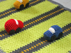Trois petites voitures au crochet — rouge, jaune et bleue — sont disposées sur un tapis de jeu crocheté vert, gris et jaune.