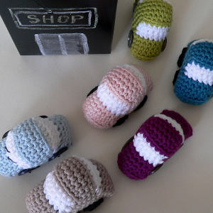 Six petites voitures jouets colorées, réalisées au crochet, sont disposées autour d'un tableau noir sur lequel est inscrit « SHOP », reproduisant ainsi une scène de parking.
