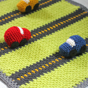 Trois petites voitures en tricot, rouge, bleue et jaune, sont posées sur un tapis de jeu tricoté vert et gris, conçu pour ressembler à une route avec des lignes jaunes en pointillés.
