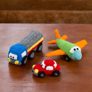 Trois jouets colorés réalisés au crochet — un camion bleu et gris, une voiture rouge et un avion vert aux ailes orange — sont posés sur un support en bois.