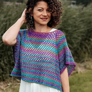 Une femme aux cheveux bouclés sourit en plein air, vêtue d'un poncho coloré au crochet, dans des tons de bleu, de violet et de rose, qu'elle porte par-dessus une robe blanche. En arrière-plan, on aperçoit du feuillage vert flou.