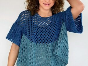 Une personne aux cheveux bouclés porte un chemisier au crochet bleu et turquoise, au motif ample et aéré ; elle sourit et prend la pose, une main posée sur la tête, devant un fond uni.