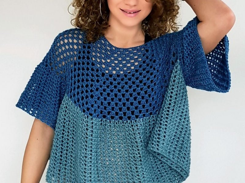 Une personne aux cheveux bouclés porte un chemisier au crochet bleu et turquoise, au motif ample et aéré ; elle sourit et prend la pose, une main posée sur la tête, devant un fond uni.