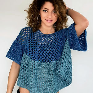 Femme aux cheveux bouclés, vêtue d'un haut bleu en crochet à manches courtes, souriant à l'appareil photo, une main posée sur ses cheveux, devant un fond uni.