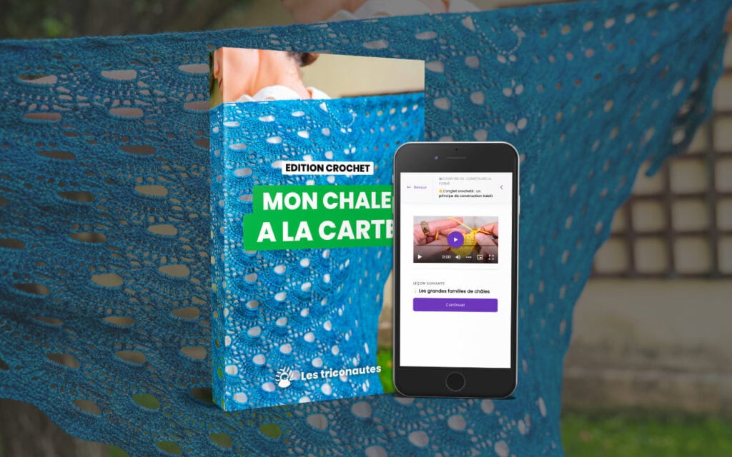 Un châle en dentelle bleue au crochet est exposé derrière un livre de modèles de crochet intitulé « Mon Châle à la Carte » et un smartphone sur lequel est diffusée une vidéo tutorielle de crochet, idéale pour tous ceux qui souhaitent perfectionner leurs compétences en matière de crochet.