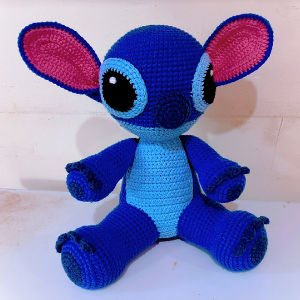 Une peluche bleue au crochet, dotée de grands yeux noirs, de grandes oreilles roses et bleues et de membres ronds, assise sur une surface blanche devant un fond clair.