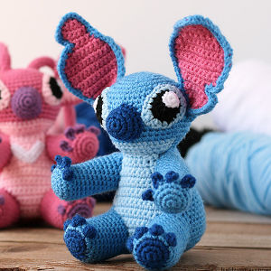 Une peluche bleue au crochet, dotée de grandes oreilles roses et bleues et de grands yeux noirs, est posée sur une surface en bois. Une peluche rose au crochet et des bobines de laine apparaissent floues à l'arrière-plan.