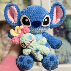 Une main tient une peluche bleue représentant Stitch, du film « Lilo & Stitch ». Stitch tient lui-même une petite peluche verte représentant une poupée vaudou, avec des yeux en boutons, une bouche cousue et un nœud rose et jaune sur la tête.
