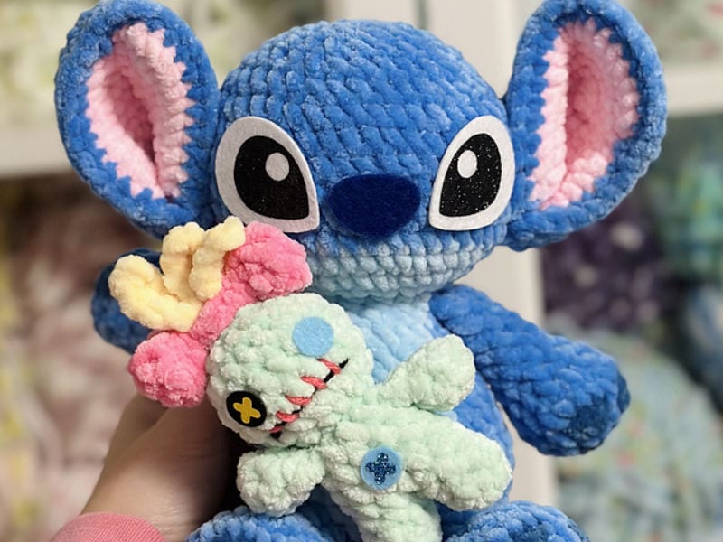 Une peluche représentant Stitch, du film « Lilo & Stitch », tient dans ses mains une petite poupée en peluche verte ornée d'un nœud rose et d'un œil en bouton, idéale pour les fans qui adorent les créations « Stitch au crochet ».