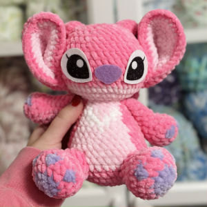 Une main tient une peluche rose et violette aux grands yeux, aux grandes oreilles et au ventre blanc. À l'arrière-plan, on aperçoit des étagères floues sur lesquelles sont disposées d'autres peluches.
