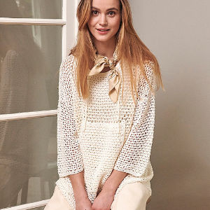 Une femme aux longs cheveux blonds, vêtue d'un haut blanc ample en crochet, d'un pantalon crème et d'une écharpe beige autour du cou, est assise à l'intérieur près d'une porte vitrée et sourit à l'appareil photo.