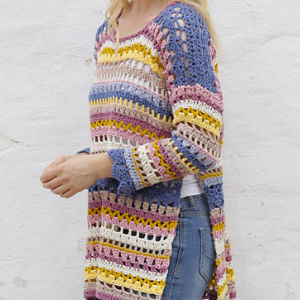 Une femme portant un pull coloré à manches longues, tricoté au crochet, avec des rayures violettes, jaunes, blanches et bleues, assorti à un jean bleu, debout devant un mur blanc.