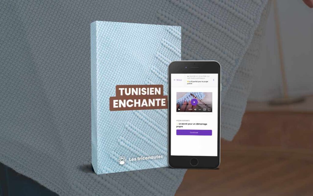 Un livre intitulé « Tunisien Enchanté » est posé à côté d'un smartphone sur lequel est diffusée une vidéo tutoriel de tricot. En arrière-plan, on aperçoit un tissu tricoté texturé, de couleur bleu clair.