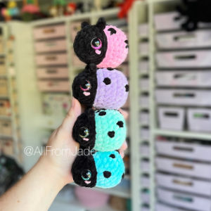 Une main tient quatre peluches de coccinelles au crochet, de couleur rose, violette, bleue et turquoise, chacune avec des taches et un visage noirs, devant un fond de tiroirs de rangement blancs.