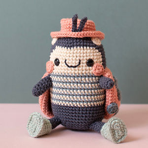 Une peluche au crochet représentant un insecte souriant, avec un corps rayé, un bonnet rose, des joues roses, des ailes et des pattes vert clair, assise sur un fond aux couleurs pastel.