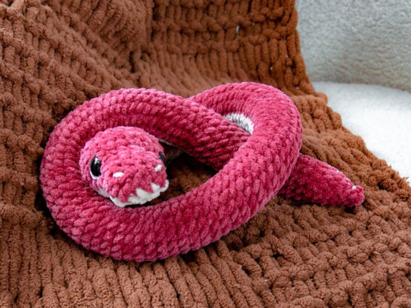 Un serpent en peluche rose, tricoté, avec des détails blancs, repose sur une couverture marron texturée, mettant en valeur le charme des tutoriels d'amigurumis au crochet.