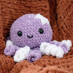 Une pieuvre en peluche violette, avec de grands yeux noirs, un petit sourire et des touches de blanc sur la tête et les tentacules, est posée sur une couverture marron texturée.