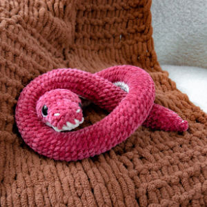 Un serpent en peluche rose, aux yeux brodés et aux dents blanches, est lové sur une couverture marron texturée.