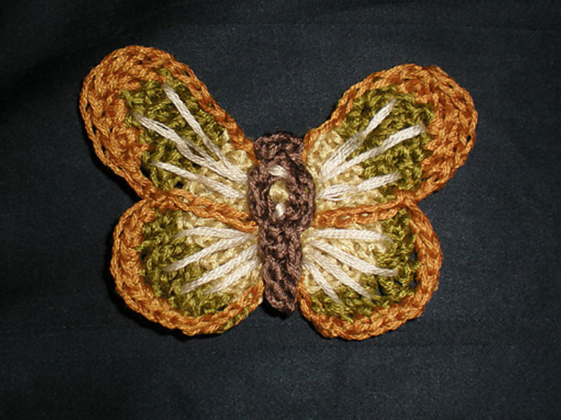 Un papillon au crochet, réalisé avec du fil marron, vert, beige et orange, repose sur un fond de tissu foncé. Ses ailes présentent des couleurs superposées et un motif texturé, ce qui en fait un exemple charmant pour ceux qui recherchent des tutoriels d’appliqués d’animaux au crochet.
