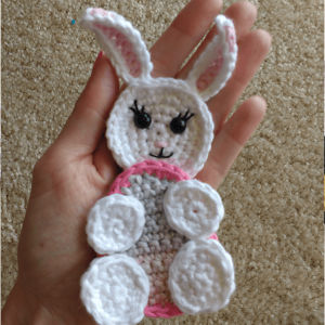 Une main tient un lapin au crochet, réalisé avec du fil blanc et rose, doté d'un visage souriant, de longues oreilles et de pattes rondes, sur un fond de tapis beige.
