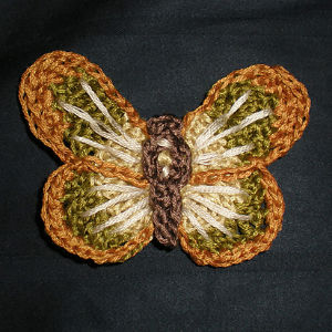 Un papillon au crochet, dont les ailes sont réalisées en fil marron, beige et vert et le corps en fil marron foncé, présenté sur un fond noir.