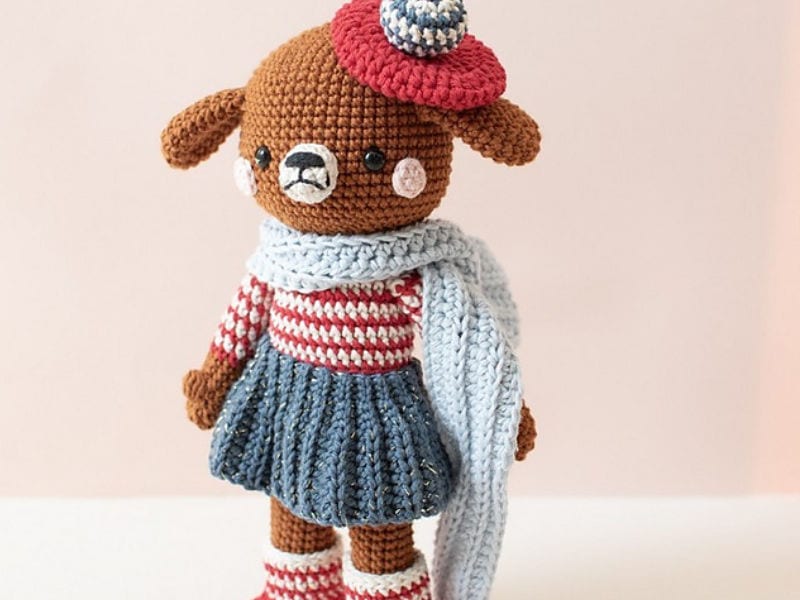 Ours en peluche au crochet vêtu d’un haut rayé rouge et blanc, d’une jupe bleue, de chaussures rouges, d’une écharpe bleue et d’un bonnet rouge surmonté d’une boule bleue et blanche, posé devant un fond clair — il s’intègre à merveille dans n’importe quelle collection de peluches ou côtoie à merveille vos créations préférées de « chien au crochet ».