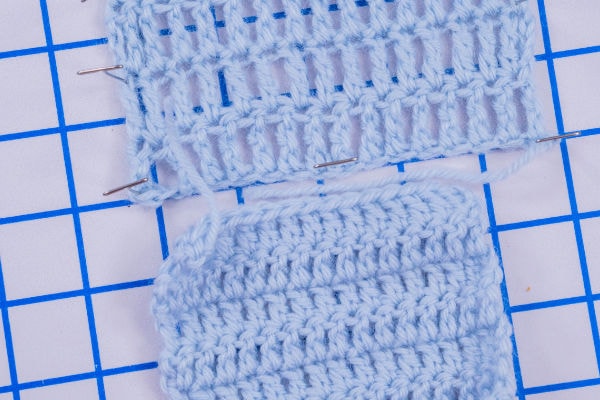 Deux échantillons de crochet bleu clair, l’un en dentelle et l’autre uni, sont disposés sur une grille bleue et blanche. L’échantillon en dentelle est fixé à l’aide d’épingles à coudre, et les deux échantillons au crochet sont réalisés avec un fil similaire.