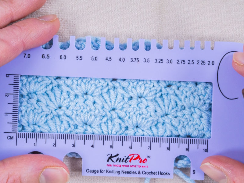 Deux mains tiennent une règle de mesure KnitPro bleue servant à mesurer un échantillon au crochet — un morceau de tissu au crochet bleu clair — sur laquelle sont indiquées les dimensions en centimètres et en millimètres.