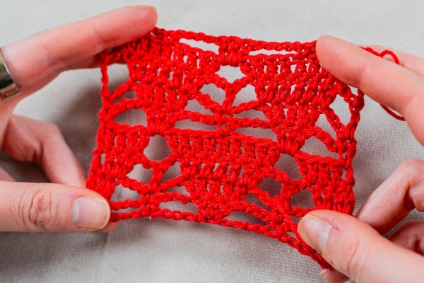 Deux mains tiennent un petit échantillon au crochet de forme rectangulaire, réalisé en dentelle rouge au crochet et orné de motifs géométriques, mis en valeur sur un fond neutre.