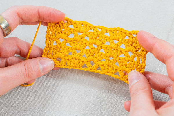 Deux mains tiennent un petit échantillon au crochet de forme rectangulaire, réalisé en fil jaune, présentant un motif ajouré et dentelé sur un fond clair.