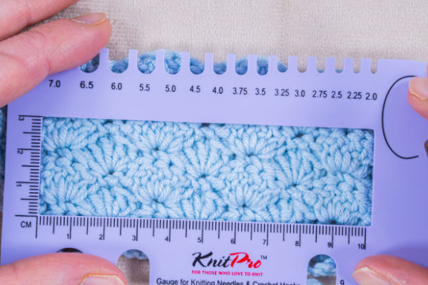 Gros plan sur des mains tenant une règle à mailles en tricot bleue au-dessus d'un échantillon au crochet bleu clair, afin de mesurer les mailles de l'échantillon. La règle comporte des graduations en centimètres et en pouces pour plus de précision.