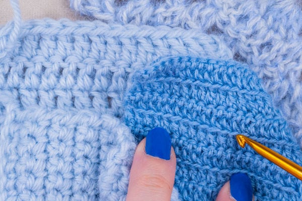 Gros plan sur des mains aux ongles vernis en bleu tenant un échantillon au crochet bleu clair, avec un crochet doré enfoncé dans le fil.