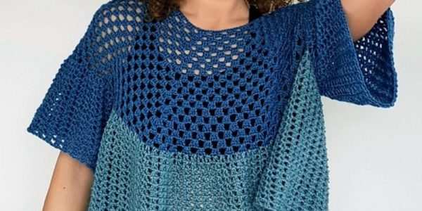 Une personne aux cheveux bouclés porte un chemisier au crochet bleu et turquoise, au motif ample et aéré ; elle sourit et prend la pose, une main posée sur la tête, devant un fond uni.