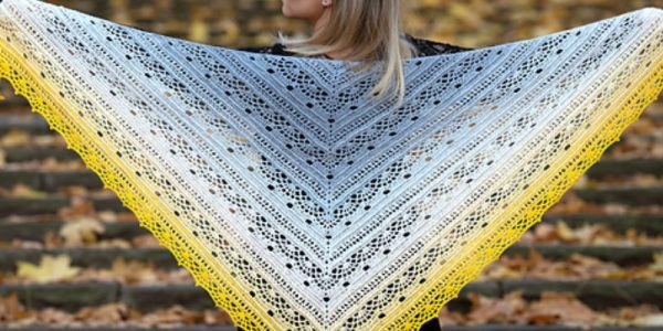 Une personne se tient dehors sur des marches en pierre recouvertes de feuilles d'automne, tenant un grand châle au crochet triangulaire aux couleurs dégradées allant du gris et blanc au centre au jaune sur les bords.