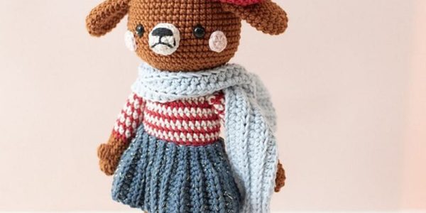 Ours en peluche au crochet vêtu d’un haut rayé rouge et blanc, d’une jupe bleue, de chaussures rouges, d’une écharpe bleue et d’un bonnet rouge surmonté d’une boule bleue et blanche, posé devant un fond clair — il s’intègre à merveille dans n’importe quelle collection de peluches ou côtoie à merveille vos créations préférées de « chien au crochet ».