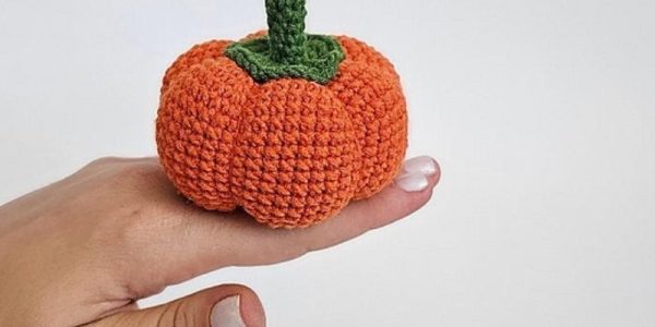 Une main aux ongles vernis de blanc tient une petite « citrouille au crochet » — une citrouille orange tricotée au crochet, dotée d'une tige verte — sur un fond blanc uni.