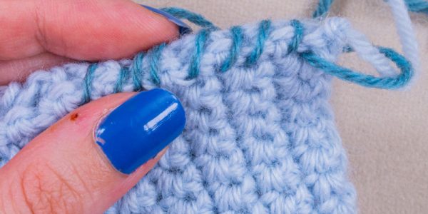 Une main aux ongles vernis de bleu tient un tissu au crochet bleu clair, tandis qu'un fil bleu-vert est cousu sur le bord supérieur, mettant en valeur un travail de haute couture au crochet d'une grande finesse.