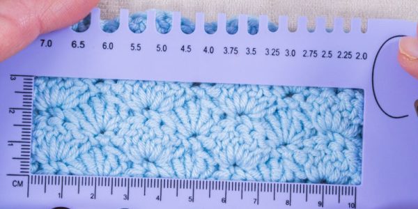 Deux mains tiennent une règle de mesure KnitPro bleue servant à mesurer un échantillon au crochet — un morceau de tissu au crochet bleu clair — sur laquelle sont indiquées les dimensions en centimètres et en millimètres.