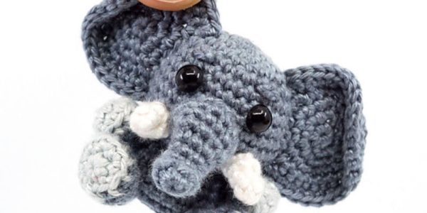 Une main brandit un petit éléphant au crochet avec de grandes oreilles, une longue trompe, des yeux noirs en forme de boutons et des défenses blanches.