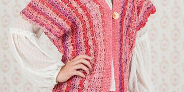 Une personne porte un gilet sans manche au crochet texturé et multicolore, rose et rouge, sur un chemisier blanc à manches bouffantes et un pantalon beige, sur un fond à motifs clairs.