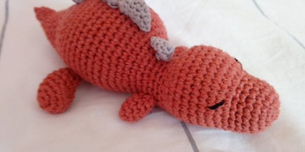 Un jouet dinosaure orange fait main au crochet, inspiré d'un modèle gratuit de dinosaure au crochet, avec les yeux fermés et trois petites pointes grises le long du dos, couché sur une couverture blanche avec de fines lignes grises.