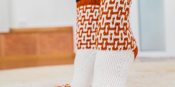 Une personne portant des chaussettes tricotées ornées d'un motif « mosaïque au crochet » orange et blanc se tient debout sur un tapis de couleur claire. Les chaussettes ont la pointe et le bord-côte orange, avec un motif répétitif sur la partie supérieure.