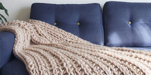 Une épaisse couverture beige en maille, confectionnée selon un motif écossais au crochet, est drapée sur un canapé bleu foncé orné de boutons. Contre un mur clair uni, une plante est partiellement visible à gauche de l'image.