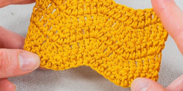 Gros plan de deux mains tenant un morceau de tissu jaune crocheté avec un motif au crochet ondulé et texturé en forme de chevron, sur un fond gris clair.